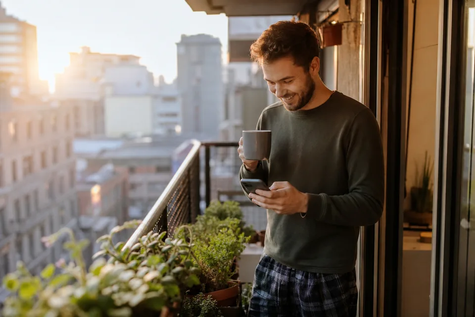 A man on a city balcony in the morning smiling at a text on his phone, coffee in hand